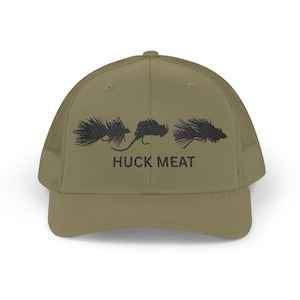 May include: Khaki green trucker cap with a mesh back and a curved peak. The front displays three black fly fishing lures above the words "HUCK MEAT".