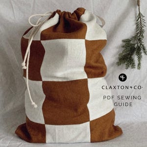 Peut inclure: Un grand sac à cordon de serrage à carreaux marron et blanc. Le sac est fait d'un tissu doux et possède une fermeture à cordon blanche. Le texte "CLAXTON + CO. PDF SEWING GUIDE" est imprimé sur le côté droit du sac.