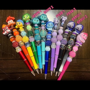May include: A set of 10 colorful pens with different characters from the movie Inside Out. Each pen has a different character on top, including Joy, Sadness, Anger, Fear, Disgust, and Ennui. The pens are decorated with beads and rhinestones.