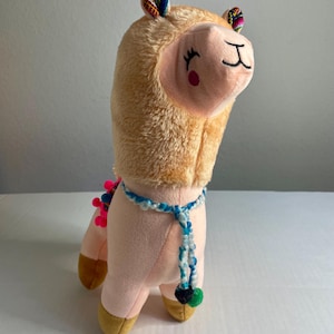 May include: A pink plush llama with a brown face, brown legs, and a blue and white beaded necklace. The llama has a colorful embroidered design on its ears and a pink pom-pom on its back.
