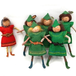 Könnte beinhalten: Eine Gruppe von sechs handgefertigten Filzpuppen. Fünf Puppen sind in grüne Filzoutfits mit braunen Gürteln und grünen Filzhüten gekleidet. Eine Puppe trägt ein rotes Filzkleid mit einem braunen Gürtel. Die Puppen haben bemalte Gesichter und Wollhaare.