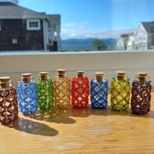 May include: Eight small glass bottles with cork tops, each covered in a colorful beaded design. The bottles are arranged in a row on a wooden surface.
