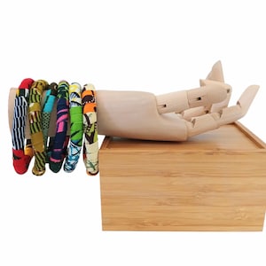May include: A collection of fabric-covered bangles in various colors and patterns, including red, yellow, green, and blue. The bracelets are displayed on a wooden hand model, resting on a small wooden box. The background is plain white.