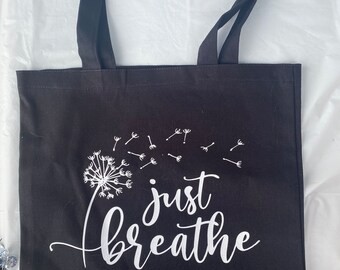 May include: A black tote bag with two shoulder straps featuring a white graphic of a dandelion with seeds blowing in the wind and the text "just breathe" written in a cursive script font centered on the front of the bag.