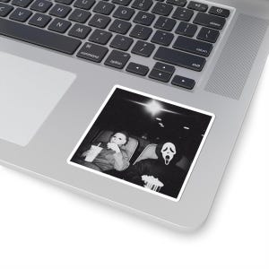 May include: Black and white sticker featuring a movie scene with two figures in costume. One figure wears a Michael Myers mask and holds a drink, while the other wears a Ghostface mask and holds popcorn. The sticker is placed on a laptop.