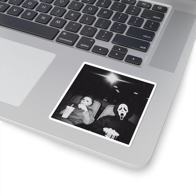 May include: Black and white sticker featuring a movie scene with two figures in costume. One figure wears a Michael Myers mask and holds a drink, while the other wears a Ghostface mask and holds popcorn. The sticker is placed on a laptop.