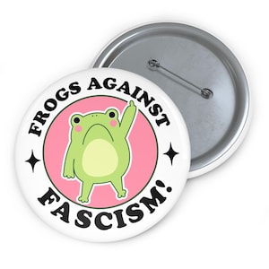 May include: A white button with the text "FROGS AGAINST FASCISM!" encircling a cartoon frog inside a pink circle. The frog is green with a white belly and is giving the middle finger. A second button is visible.