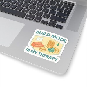 May include: A white sticker with the text "BUILD MODE IS MY THERAPY" in teal. The sticker features a cartoon illustration of a room with furniture, including a sofa, armchair, table, and bookcase. The sticker is placed on a laptop.