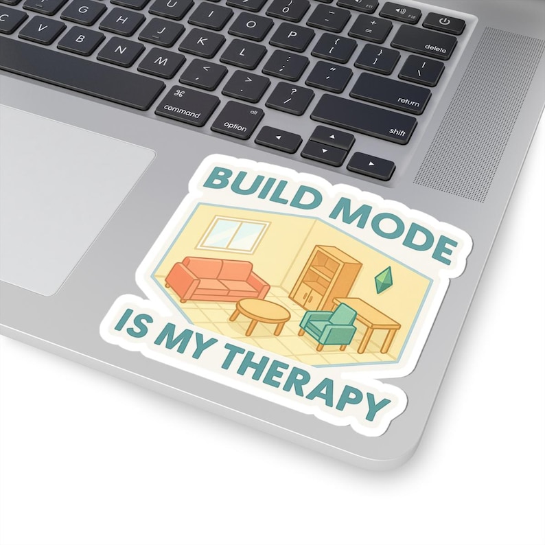 May include: A white sticker on a laptop showing a cartoon interior design scene with a red sofa, teal armchair, and wooden furniture. The text "BUILD MODE IS MY THERAPY" is displayed in teal and white.