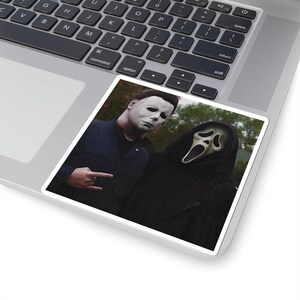 May include: A square sticker featuring a photograph of two figures in horror movie costumes. One figure wears a white mask and dark jumpsuit, while the other wears a black hooded robe and a white ghost mask. The sticker is applied to a laptop.