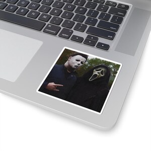 May include: A square sticker featuring a graphic of two horror movie characters: one wearing a white mask and dark blue jacket, and the other wearing a black hooded robe with a white ghost mask. The sticker is placed on a silver laptop keyboard.