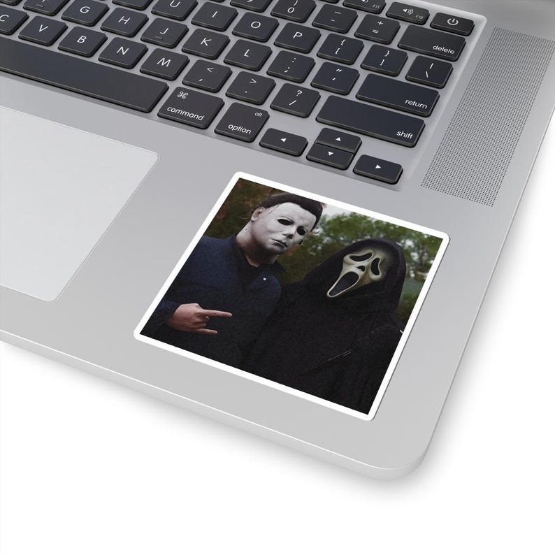 May include: A square sticker featuring a graphic of two horror movie characters: one wearing a white mask and dark blue jacket, and the other wearing a black hooded robe with a white ghost mask. The sticker is placed on a silver laptop keyboard.