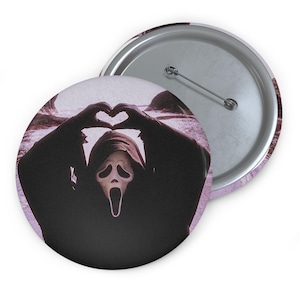 May include: A round pin-back button featuring a graphic of a hooded figure with a white ghost-like mask and a black robe. The figure is making a heart shape with its hands above its head. The background is a muted purple landscape.