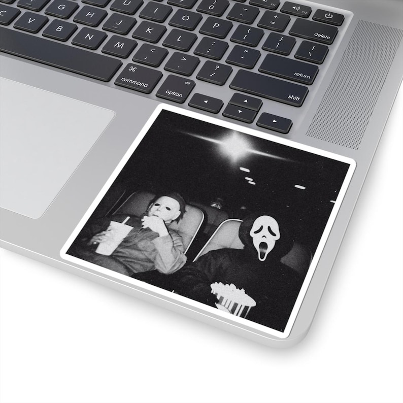 May include: Black and white sticker featuring a movie scene with two characters in costume. One character is wearing a white mask and holding a drink. The other character is wearing a Scream mask and holding popcorn.