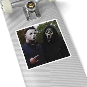 May include: A skateboard deck featuring a square sticker with a graphic of Michael Myers in a dark blue shirt and the Ghostface character in a black hooded robe. The skateboard has a light gray and white diagonal striped pattern and silver trucks.