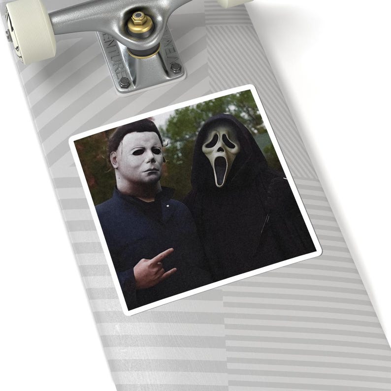 May include: A skateboard deck featuring a square sticker with a graphic of Michael Myers in a dark blue shirt and the Ghostface character in a black hooded robe. The skateboard has a light gray and white diagonal striped pattern and silver trucks.