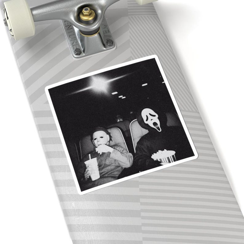 May include: Black and white sticker featuring a movie theater scene with two figures in costume. One figure wears a Michael Myers mask and holds a drink, while the other wears a Ghostface mask and holds popcorn. The sticker is placed on a skateboard.