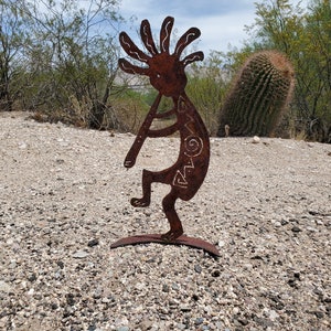 May include: A rusty metal Kokopelli figure playing a flute, standing on a curved base. The figure has a stylized design with a curved body, a head with radiating lines, and decorative markings. The sculpture is outdoors, set against a backdrop of desert vegetation.