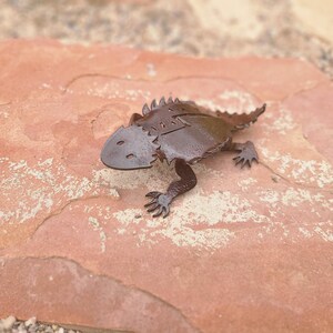 May include: A metal horned lizard sculpture with a detailed design. The lizard is dark brown and has a textured appearance. It is resting on a reddish-brown stone surface.