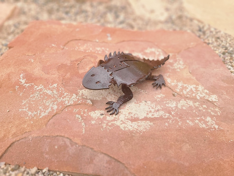 May include: A metal horned lizard sculpture with a detailed design. The lizard is dark brown and has a textured appearance. It is resting on a reddish-brown stone surface.