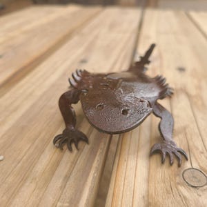 May include: A detailed, metal sculpture of a lizard with a textured, dark brown finish. The lizard has a wide head, four legs with clawed feet, and a spiky tail. The sculpture is placed on a wooden surface.