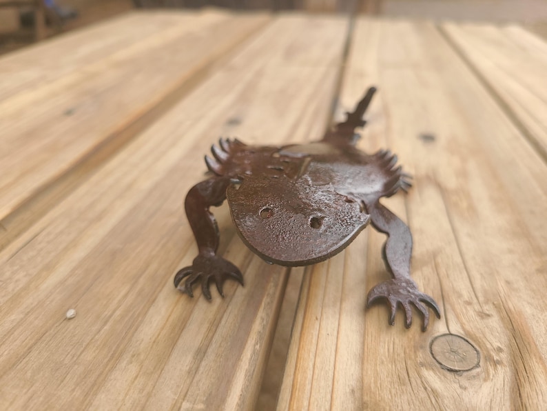 May include: A detailed, metal sculpture of a lizard with a textured, dark brown finish. The lizard has a wide head, four legs with clawed feet, and a spiky tail. The sculpture is placed on a wooden surface.