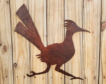 May include: A rusty metal roadrunner silhouette, mounted on a light-colored wooden plank background. The bird is depicted in mid-stride, with its long tail feathers extended. This rustic wall art adds a touch of desert charm.