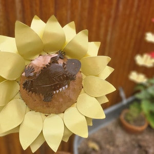 May include: A metal sculpture of a sunflower with a brown and tan platypus in the center. The sunflower petals are a pale yellow color. The background is blurred, with a glimpse of a garden.