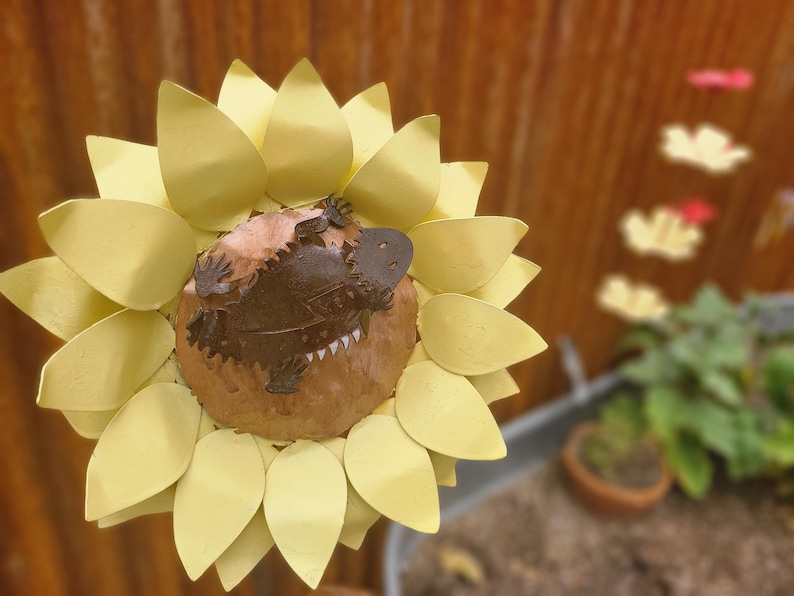 May include: A metal sculpture of a sunflower with a brown and tan platypus in the center. The sunflower petals are a pale yellow color. The background is blurred, with a glimpse of a garden.
