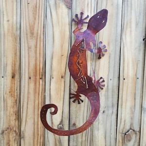May include: A rusty metal gecko sculpture with intricate cut-out designs, mounted on a wooden fence. The lizard has a curled tail and detailed feet, showcasing a blend of orange, purple, and brown hues. This decorative piece adds a rustic touch to outdoor spaces.
