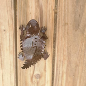 May include: A metal lizard-shaped wall decoration with a lightning bolt design. The lizard is brown and has a textured surface. It is mounted on a wooden background.