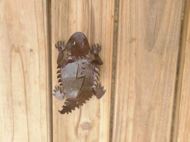 May include: A metal lizard-shaped wall decoration with a lightning bolt design. The lizard is brown and has a textured surface. It is mounted on a wooden background.