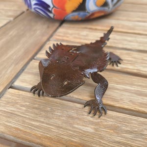May include: A metal sculpture of a lizard, featuring a detailed design with a textured surface and spiky protrusions. The lizard is brown and rests on a wooden surface. A decorative ceramic bowl with colorful patterns is in the background.