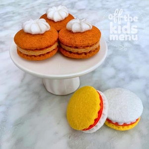 Op de afbeelding: Vilt speelgoed set met drie oranje vilt sandwiches met witte slagroom toppings op een witte taartstandaard. Twee extra vilt sandwiches, een gele en een witte, zijn ook zichtbaar.