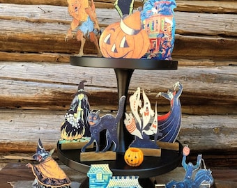 May include: A tiered cake stand with a black base and wooden tiers, decorated with vintage Halloween cutouts. The cutouts include a witch, a werewolf, a skeleton, a vampire, a ghost, a haunted house, a jack-o-lantern, and a black cat. There are also small orange pumpkins on the stand.