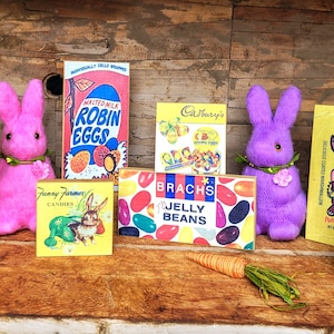 May include: A collection of vintage Easter candy boxes featuring brands like Reese's, Brach's, Robin Eggs, and Cadbury's. The boxes are arranged on a wooden surface with two plush pink and purple bunnies.