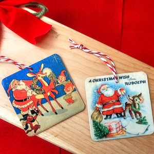 May include: Two vintage Christmas ornaments featuring Santa Claus and Rudolph the Red-Nosed Reindeer. The ornaments are square and have a blue background with white trim. The ornaments are tied with red and white twine.