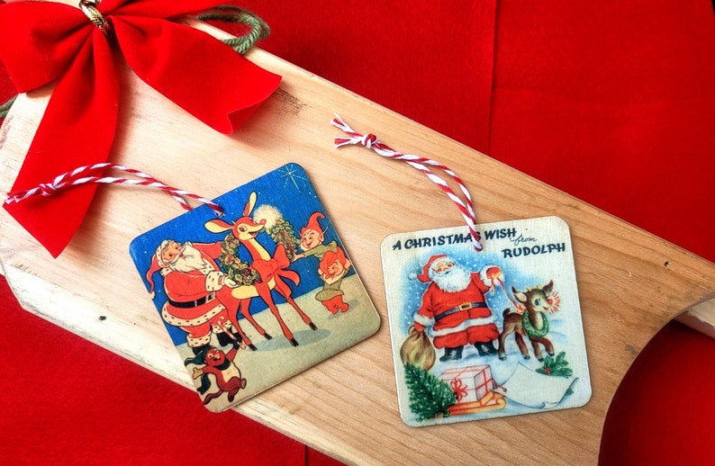 May include: Two vintage Christmas ornaments featuring Santa Claus and Rudolph the Red-Nosed Reindeer. The ornaments are square and have a blue background with white trim. The ornaments are tied with red and white twine.