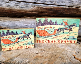 May include: Two wooden signs with a vintage Christmas scene featuring a sleigh full of presents pulled by a horse with a cowboy riding. The signs have the text "Merry Christmas Y'all" and the family names "The Ballard Family" and "The Chavis Family".