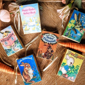 May include: Six miniature wooden books with colorful illustrations and titles. The books are "The Golden Egg Book", "Happy Easter, Mother Duck", "Here Comes Peter Cottontail", "The Adventures of Peter Cottontail", "Little Cottontail", and "Peter Rabbit".