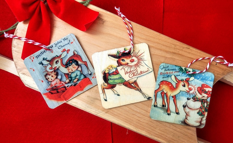 May include: Three vintage Christmas ornaments featuring Rudolph the Red-Nosed Reindeer. The ornaments are made of wood and have a rustic look. The first ornament features Rudolph with a red nose and antlers, pulling a sleigh with a green bag that says "Rudolph". The second ornament features Rudolph with a red nose and antlers, playing music with two children. The third ornament features Rudolph with a red nose and antlers, standing next to a snowman.
