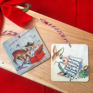 May include: Two wooden Christmas ornaments featuring a cartoon reindeer. One ornament shows the reindeer pulling a red sled with the text "Listen to Rudolph's Christmas Song". The other ornament shows the reindeer writing a letter with the text "Dear Mummy and Daddy, I've gone to help Santa. Don't worry, Rudolph."
