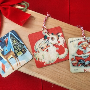 May include: Three vintage Christmas ornaments with red and white string hangers. The ornaments are square and made of wood. The first ornament features a reindeer standing in front of a toy store. The second ornament features Santa Claus with a reindeer. The third ornament features Santa Claus reading a book with a reindeer. The text on the third ornament reads "Looks like it's Christmas, says Rudolph".
