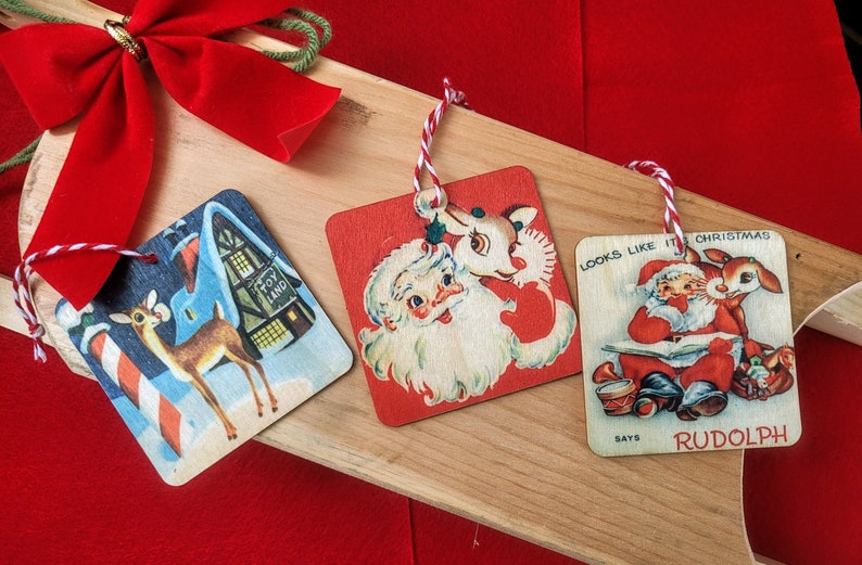 May include: Three vintage Christmas ornaments with red and white string hangers. The ornaments are square and made of wood. The first ornament features a reindeer standing in front of a toy store. The second ornament features Santa Claus with a reindeer. The third ornament features Santa Claus reading a book with a reindeer. The text on the third ornament reads "Looks like it's Christmas, says Rudolph".