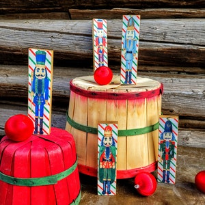 May include: Five wooden nutcracker ornaments with different designs. The nutcrackers are painted in red, blue, green, and gold. They are arranged on a wooden barrel and a red basket.