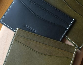 May include: Three rectangular card holders in black and olive green, each featuring white contrast stitching along the edges and a curved pocket design. The word ALDER is embossed in a clean, sans-serif font on the front of each card holder.