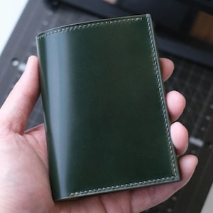 May include: A dark forest green leather card holder or wallet featuring a smooth, polished finish and visible tonal stitching along the edges. The rectangular accessory has a clean, structured design with rounded corners and a uniform surface.