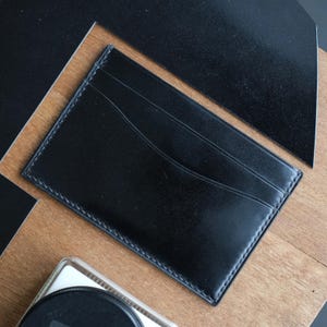 Può includere: Un porta carte in pelle nera di forma rettangolare con angoli arrotondati e cuciture visibili lungo i bordi. La superficie anteriore presenta un'apertura curva progettata per contenere carte, con una finitura liscia e opaca.