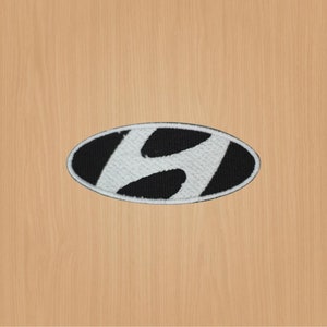 May include: An embroidered patch featuring the Hyundai logo. The logo is an oval shape with a black background and a white, stylized "H" in the center. The patch has a white border and is set against a light brown wooden surface.