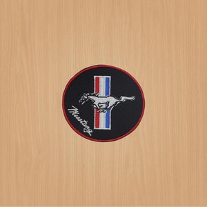 Puede incluir: Un parche bordado en blanco y negro con un caballo corriendo y la palabra "Mustang" en letras blancas. El caballo corre sobre una franja roja, blanca y azul.